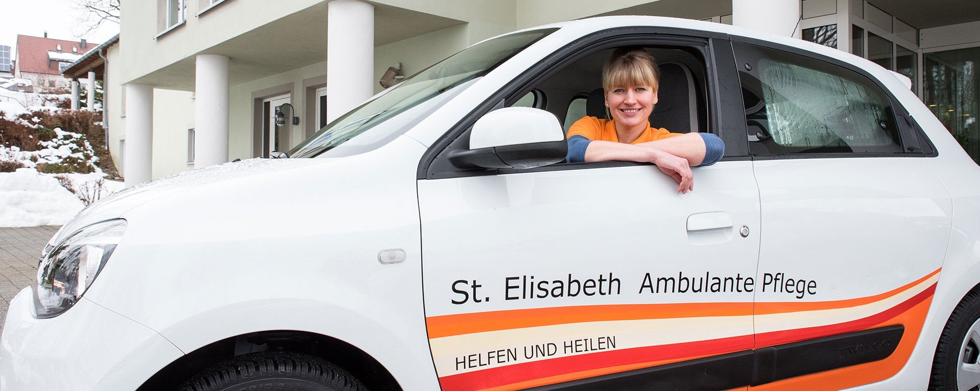 Eine junge Pflegerin in ihrem Auto mit der Aufschrift: "St. Elisabeth Ambulante Pflege - Heilen und Helfen"