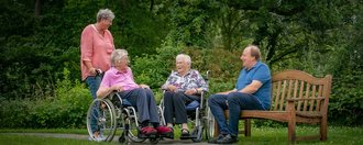 Im Garten: Ein Mann sitzt auf einer Parkbank und unterhält sich mit zwei Frauen im Rollstuhl - eine weitere Frau hält einen Rollstuhl