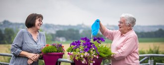 Zwei Frauen stehen an einem Geländer und gießen Blumen - lachen dabei