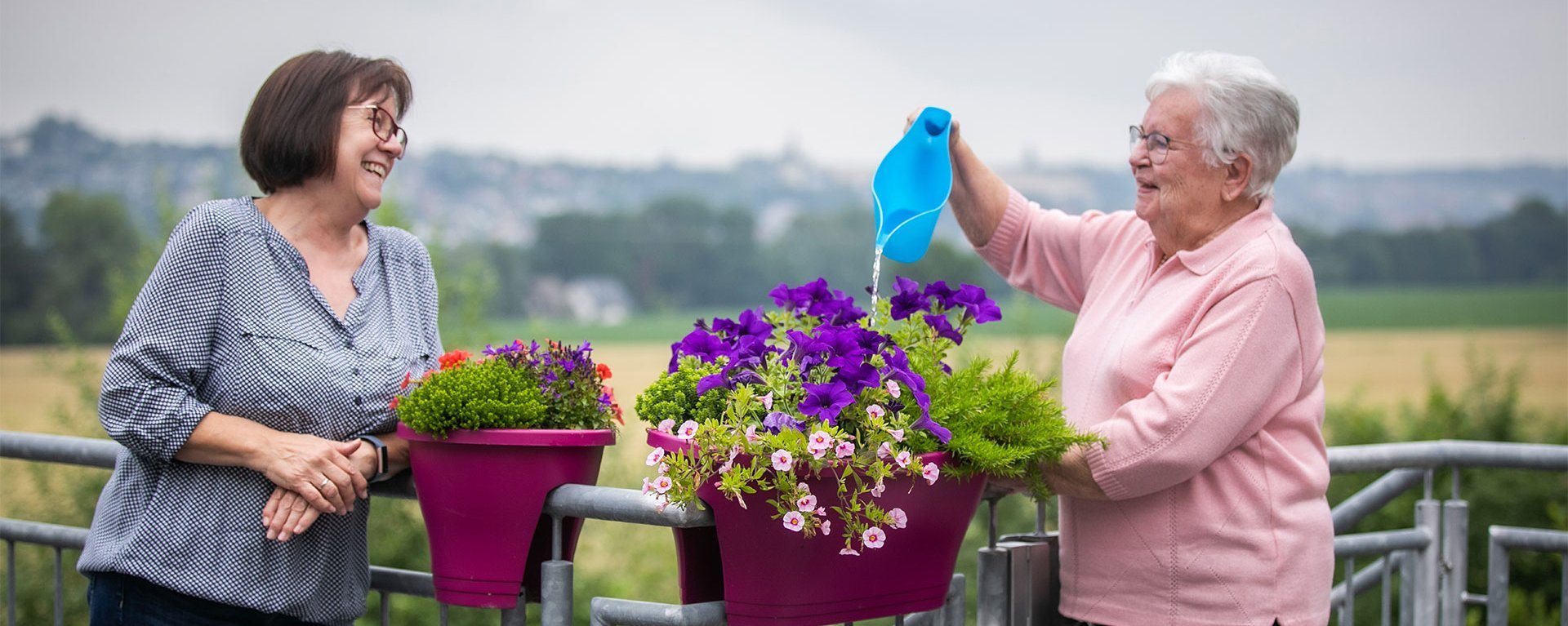 Zwei Frauen stehen an einem Geländer und gießen Blumen - lachen dabei